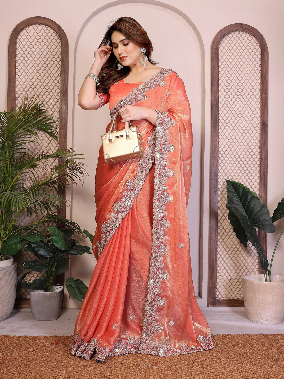 Woman in an orange saree with silver border holding a gold handbag indoors.