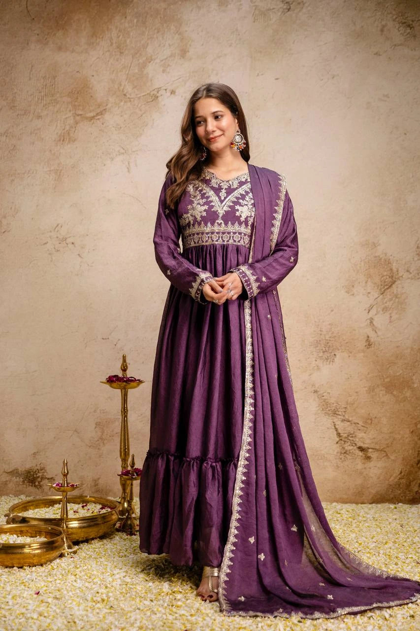 Woman in a purple traditional outfit standing against a textured beige wall.