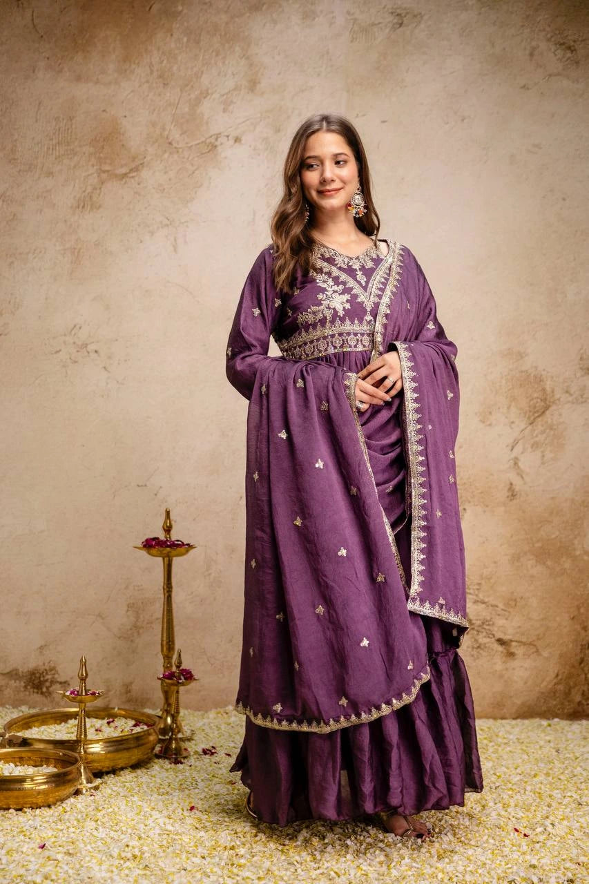 Woman in a purple traditional outfit with intricate designs, standing against a textured beige wall.
