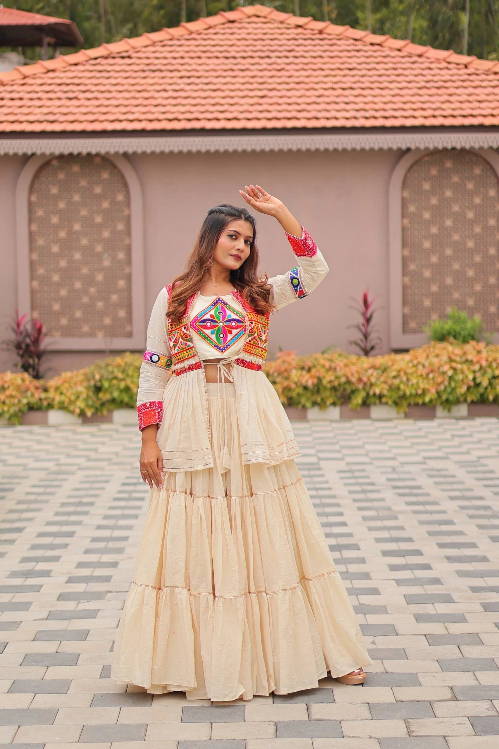 Woman in traditional outfit standing on a paved area with a building and plants in the background