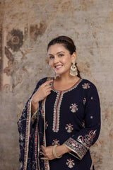 Woman wearing a navy blue embroidered traditional outfit against a textured wall.