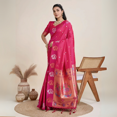 A woman models a luxurious Deep Fuchsia Soft Silk Saree. The saree features silver Zari motifs on the body and a wide pallu in contrasting rose gold, richly woven with multi-colored peacock and floral designs. She wears a matching short-sleeve pink blouse and heavy gold Kundan jewelry, posing indoors.