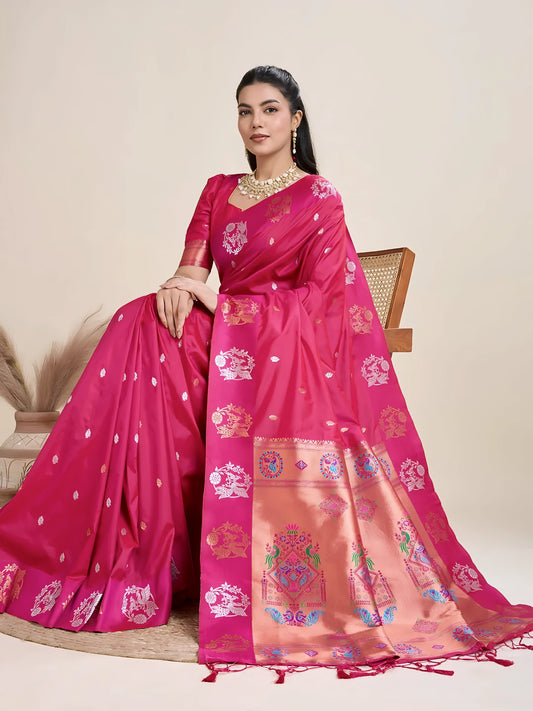 A woman models a luxurious Deep Fuchsia Soft Silk Saree. The saree features silver Zari motifs on the body and a wide pallu in contrasting rose gold, richly woven with multi-colored peacock and floral designs. She wears a matching short-sleeve pink blouse and heavy gold Kundan jewelry, posing indoors.