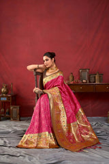 A woman models a luxurious Hot Pink Soft Banarasi Silk Saree. The saree features dense, all-over tone-on-tone Zari weaving. The wide border and pallu showcase heavy gold Zari work with vibrant green and yellow accents. She wears a contrasting dark (black/brown) sleeveless blouse and a heavy gold Kundan choker necklace, posing indoors.