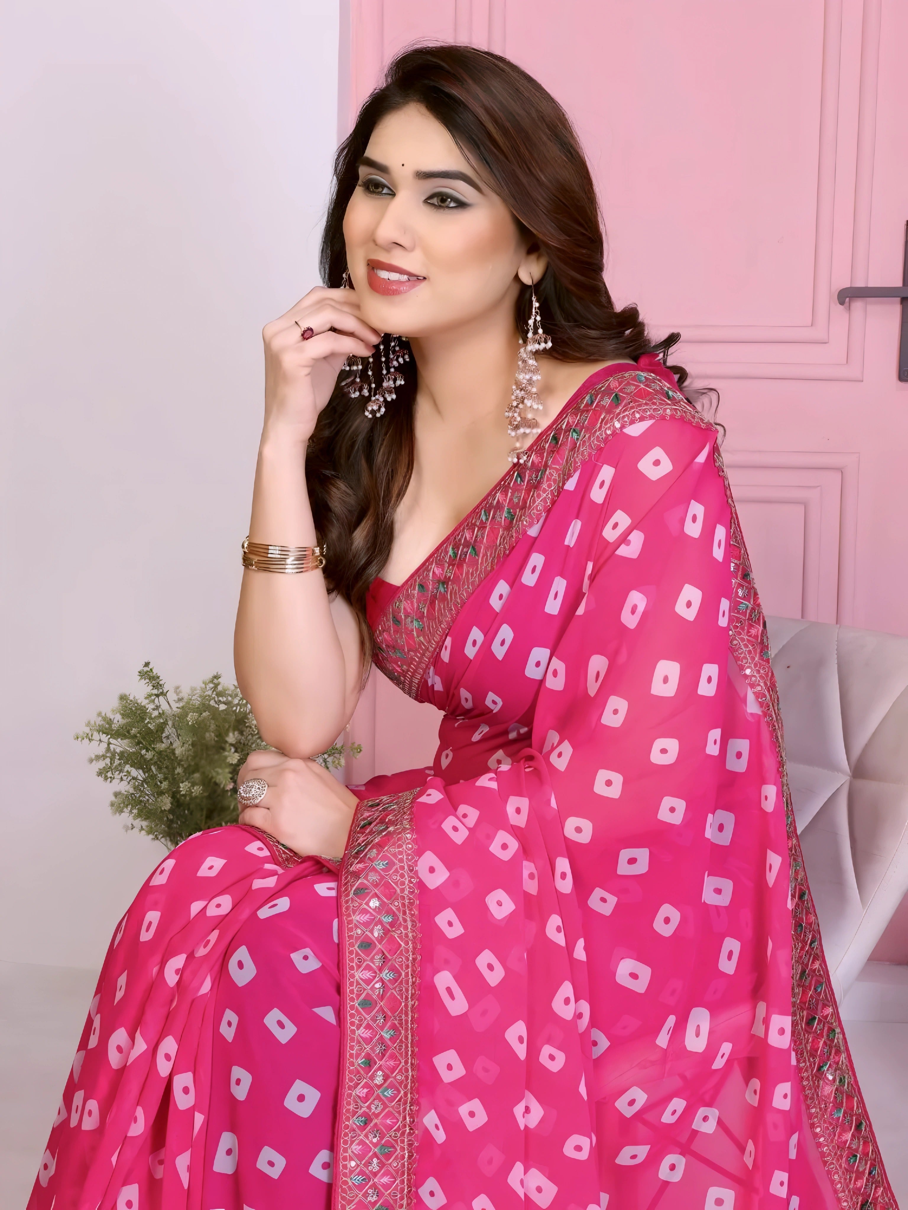Woman wearing a vibrant pink bandhani saree with traditional white tie-dye patterns and a decorative embroidered border, paired with a sleeveless matching blouse. She stands gracefully in front of a pastel pink arched doorway, accessorized with statement earrings and bangles, surrounded by earthy clay pots, a small green foliage vase, and pink decorative branches in a contemporary minimalist studio setting.