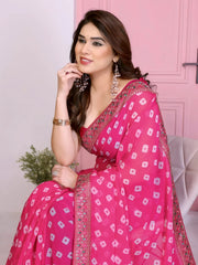Woman wearing a vibrant pink bandhani saree with traditional white tie-dye patterns and a decorative embroidered border, paired with a sleeveless matching blouse. She stands gracefully in front of a pastel pink arched doorway, accessorized with statement earrings and bangles, surrounded by earthy clay pots, a small green foliage vase, and pink decorative branches in a contemporary minimalist studio setting.