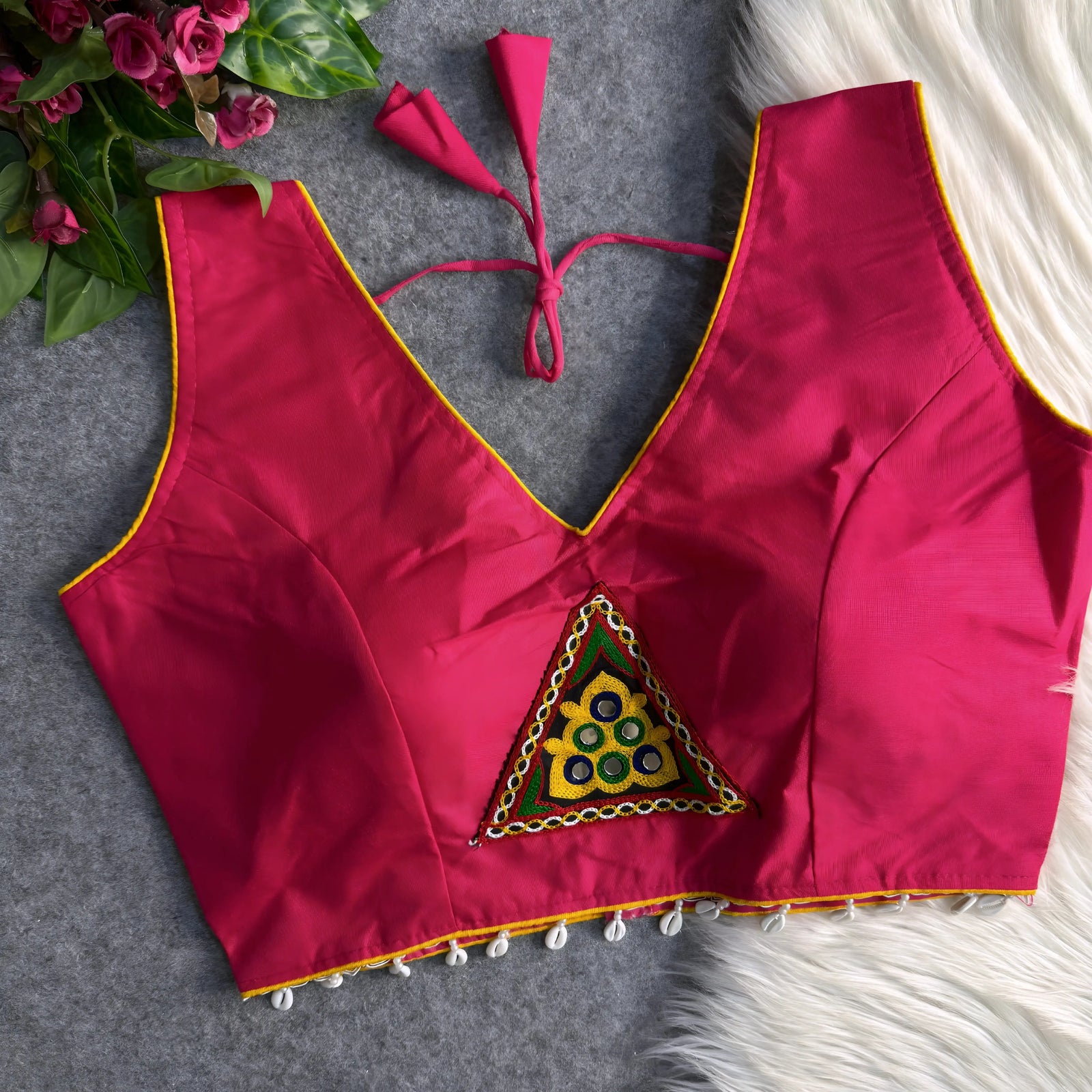 Pink padded blouse with yellow piping, featuring a central embroidered triangle motif with mirror work and shell detailing along the hem, displayed on a grey surface with artificial pink flowers and white fur in the background.