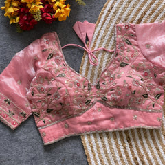  Pink padded blouse with intricate floral embroidery in gold, silver, and red threads, featuring a tie-back detail, displayed on a grey surface with a woven mat and artificial yellow and red flowers in the background.