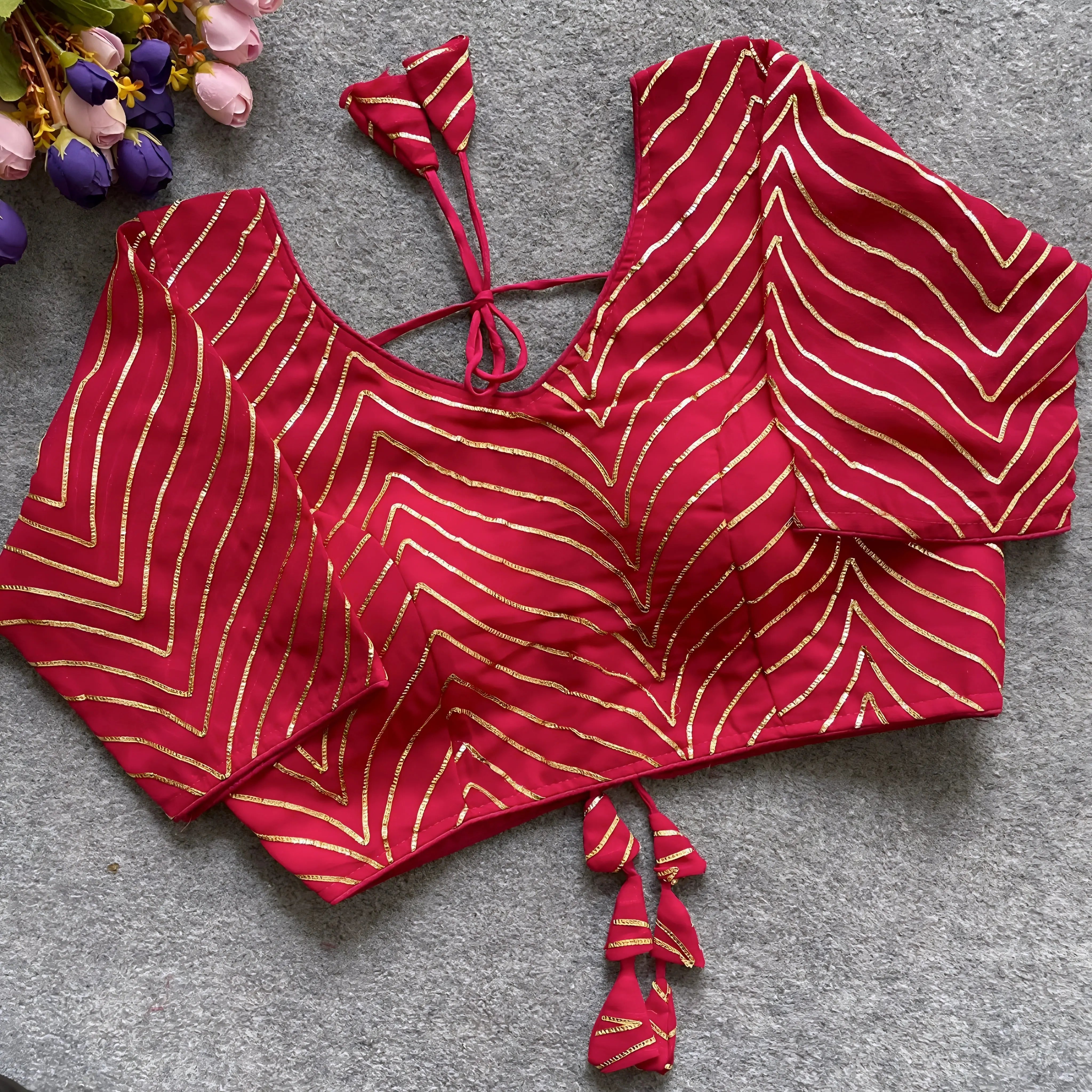 Pink blouse featuring striking gold gota patti zigzag embroidery, three-quarter sleeves, and a stylish back tie-up with matching triangular tassels. Displayed flat on a textured grey surface beside fresh pink and purple flowers, emphasizing its festive and elegant ethnic appeal.