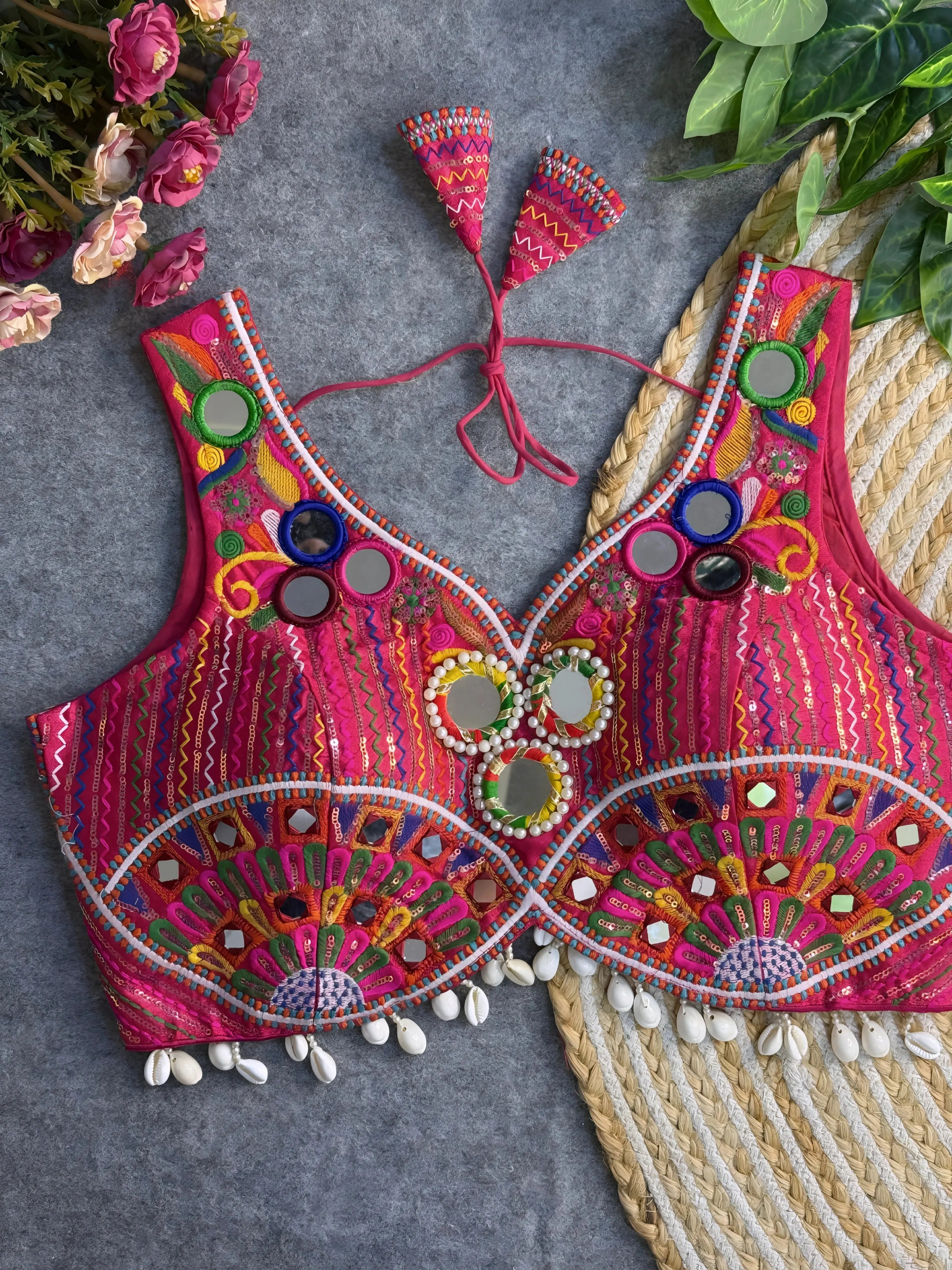Traditional pink embroidered blouse featuring vibrant multicolor mirror work, intricate thread embroidery in geometric and floral patterns, and shell tassel detailing along the hem, displayed on a flat surface with floral and leafy decor.
