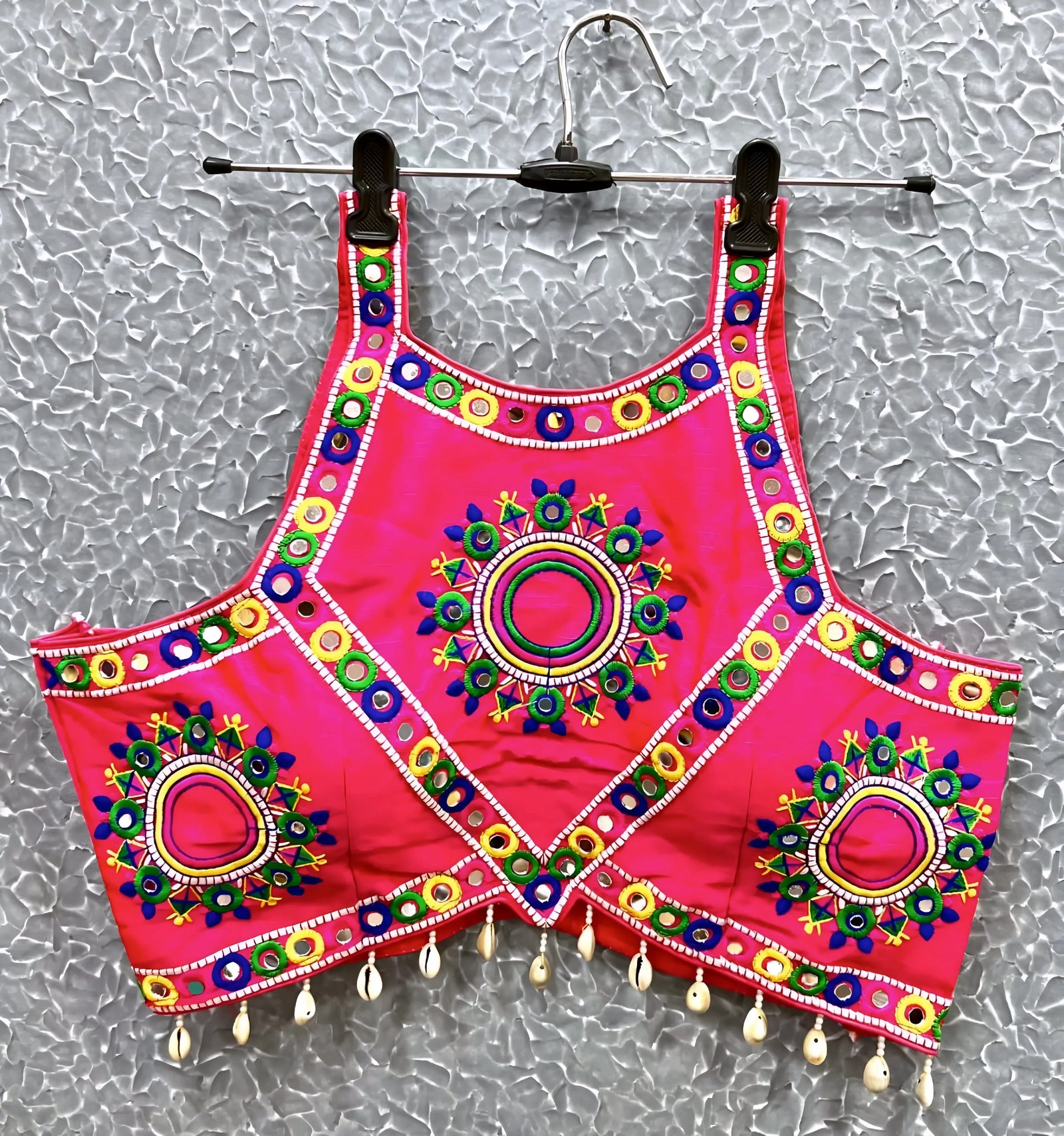 Pink blouse featuring vibrant circular and oval mirror work with traditional Kutch multicolor embroidery, adorned with geometric borders and shell tassel drops along the hem. Displayed on a hanger against a textured silver background, highlighting its festive and culturally inspired ethnic design.