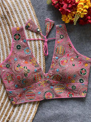 Traditional pink blouse featuring vibrant multicolor thread embroidery and mirror work in circular floral and geometric patterns, with a deep V-neck and tie-back detail, displayed flat on a textured mat with artificial red and yellow flowers in the corner.