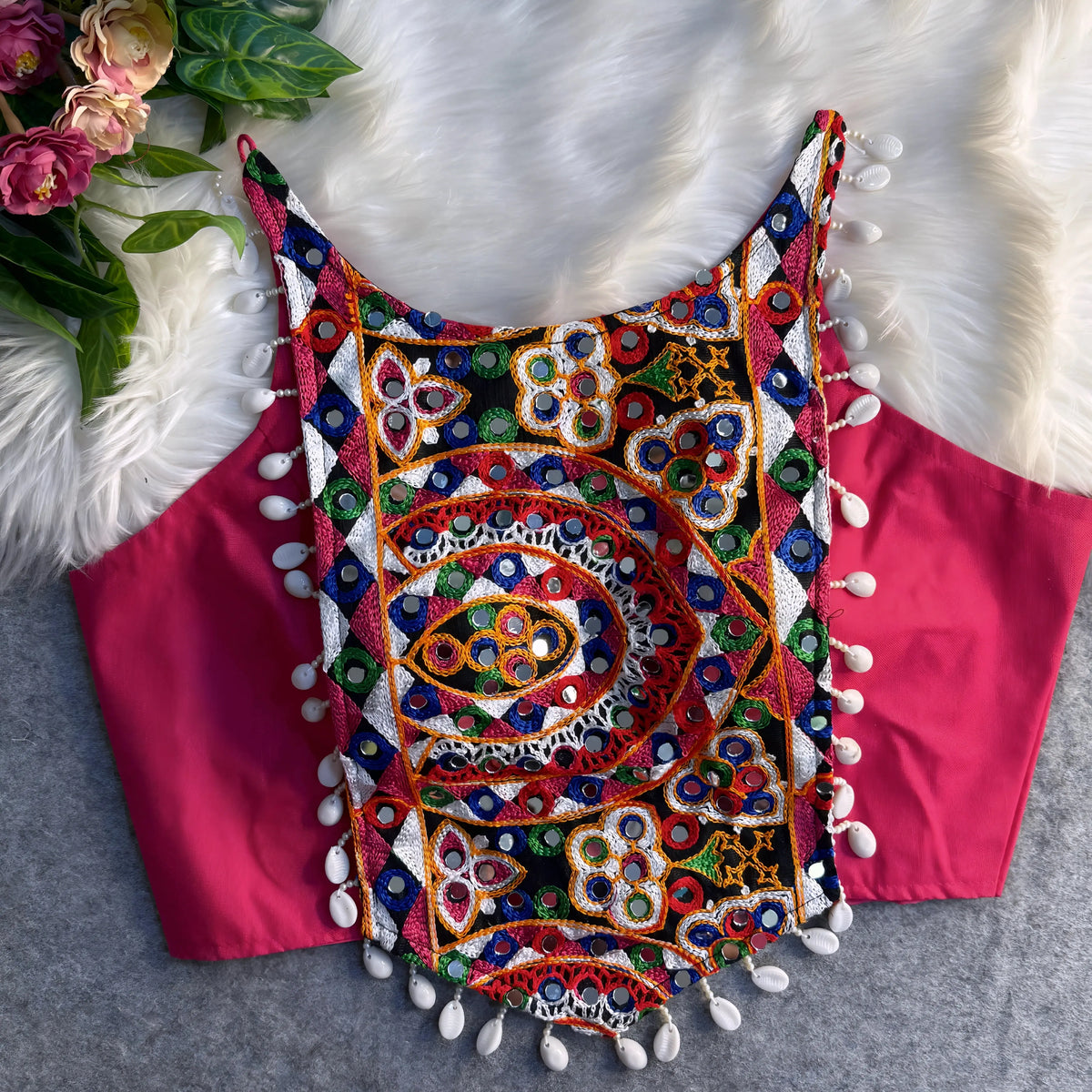 Traditional pink blouse featuring vibrant multicolor mirror work and intricate thread embroidery in geometric and floral patterns, accented with white shell tassels along the hem, displayed flat on a soft fur and grey background with floral and leafy decor.