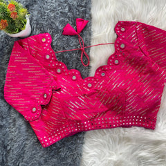 Bright pink bridal blouse with scallop sleeves, featuring circular mirror work and silver sequin stripes on the fabric, decorative borders on sleeves and hem, and tie-up tassels, displayed on a textured grey and white fur background with a small potted plant nearby.