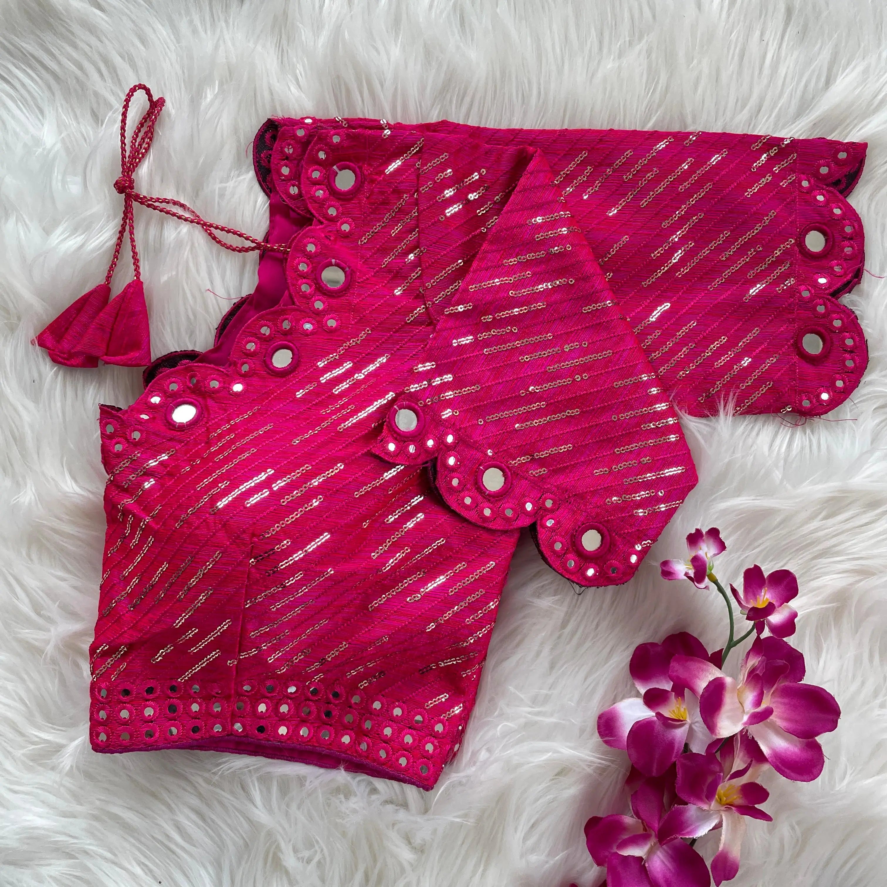 Bright pink bridal blouse with scallop sleeves, featuring circular mirror work and silver sequin stripes on the fabric, decorative borders on sleeves and hem, and tie-up tassels, displayed on a textured grey and white fur background with a small potted plant nearby.