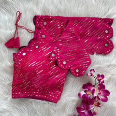 Bright pink bridal blouse with scallop sleeves, featuring circular mirror work and silver sequin stripes on the fabric, decorative borders on sleeves and hem, and tie-up tassels, displayed on a textured grey and white fur background with a small potted plant nearby.