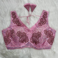 Light pink blouse featuring intricate shell-shaped maroon embroidery, sequin and bead embellishments, floral motifs, and a decorated border, with a deep V-neck and pleated fan-shaped tasseled tie-back detail, displayed flat on a white fur background.