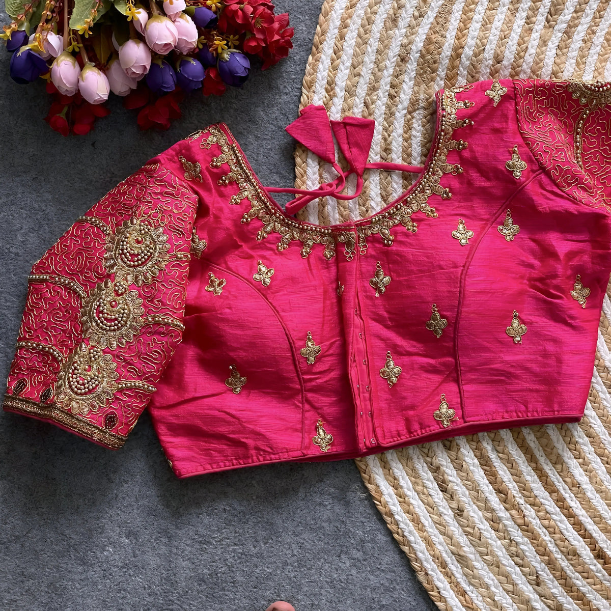 Traditional pink silk blouse featuring intricate gold embroidery and beadwork with floral and paisley motifs on the sleeves and neckline, displayed flat on a textured surface with a bunch of artificial flowers and a woven mat in the background.