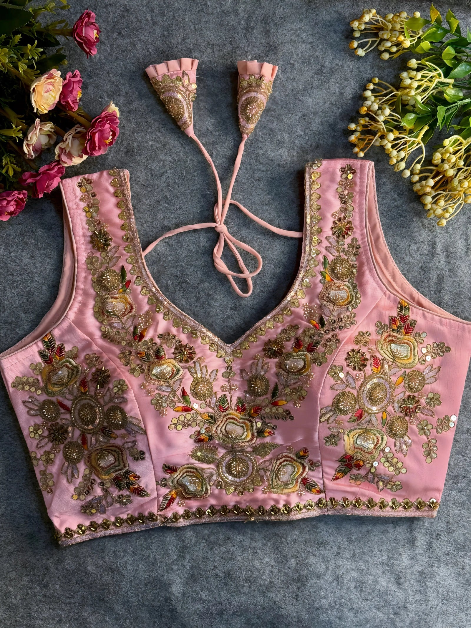 Traditional pink silk blouse featuring intricate gold zardozi embroidery and beadwork with multicolor floral and leaf motifs on the front and neckline, finished with decorative tassel ties, displayed flat on a textured grey background with artificial pink and yellow flowers in the corners.

Related
File name: vibrant-black-red-shibori-saree-tassels
Alt text: Hand-dyed black and red shibori saree with intricate tassels.