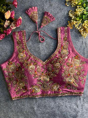 Traditional mauve silk blouse featuring intricate gold zardozi embroidery and beadwork with multicolor floral and leaf motifs on the front and neckline, finished with decorative tassel ties, displayed flat on a textured grey background with artificial pink and yellow flowers in the corners.