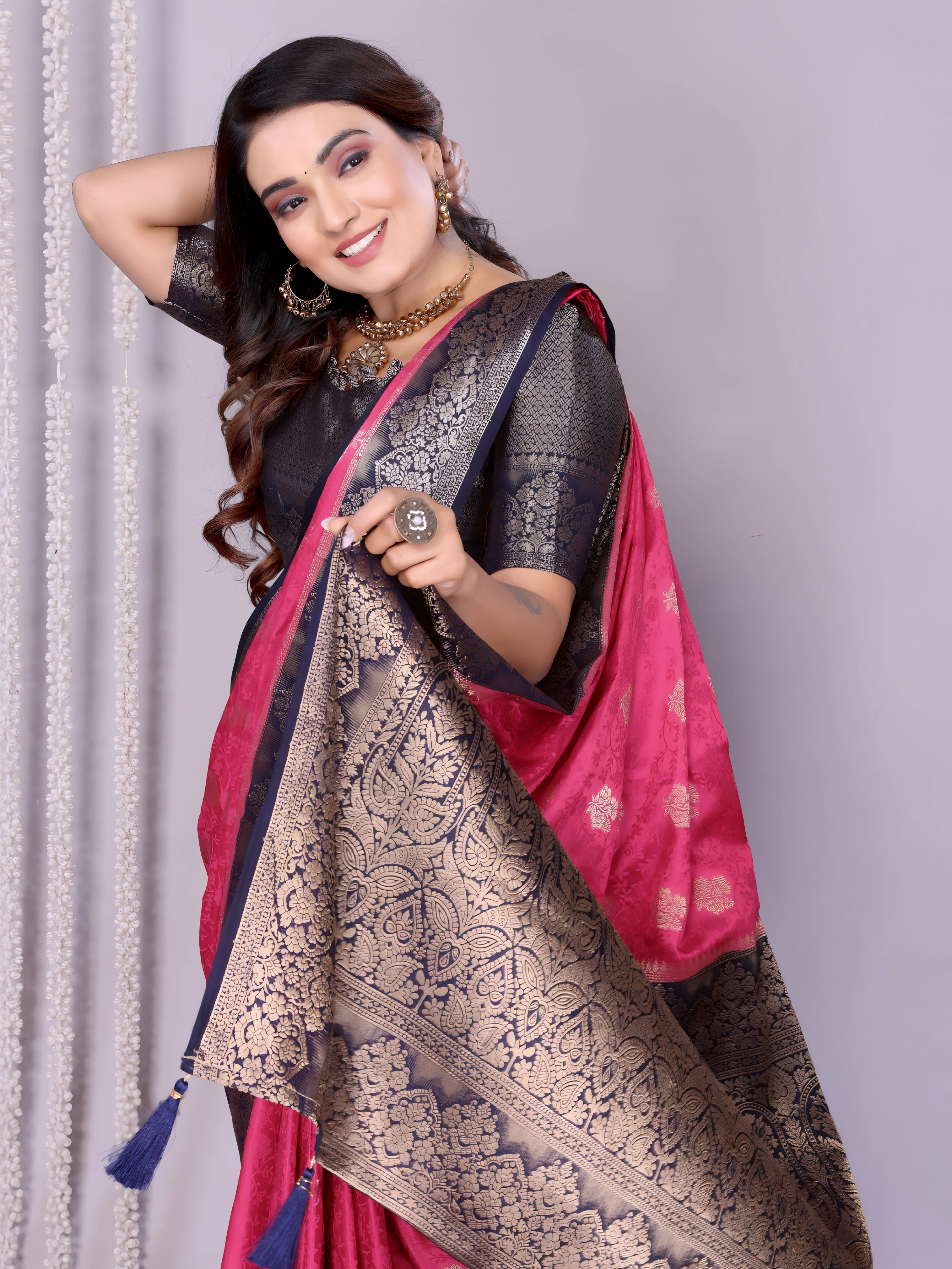 Woman wearing an elegant pink silk saree with a striking black and silver zari border and intricate woven motifs, paired with a black blouse and accessorized with traditional jewelry. She poses gracefully in a modern studio with woven baskets of rose petals and a neutral background, complemented by sheer white curtains for a refined, festive look.