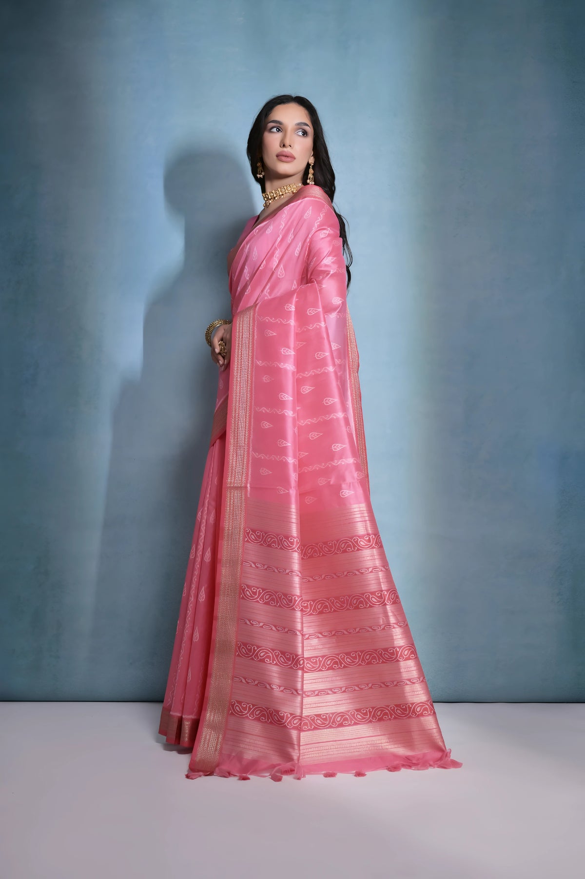 A woman models a striking Soft Pink Raw Silk Saree. The saree body features subtle woven motifs and is framed by a wide metallic gold Zari border. The pallu features intricate horizontal bands of pink and gold Ikat-style geometric weaving. She wears a matching short-sleeve pink blouse and accessorizes with a gold choker necklace and earrings, posing indoors.