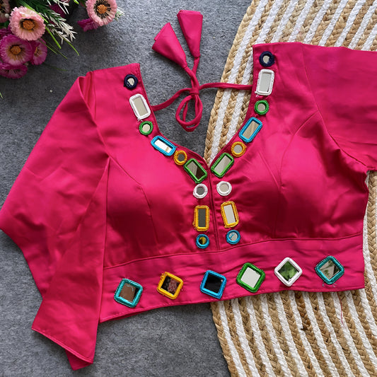 Pink blouse adorned with square and round mirror work featuring multicolored embroidered borders, stylish back tie-up details, and three-quarter sleeves. Displayed flat on a textured grey surface with woven mat and fresh pink flowers, highlighting its festive and modern ethnic design.