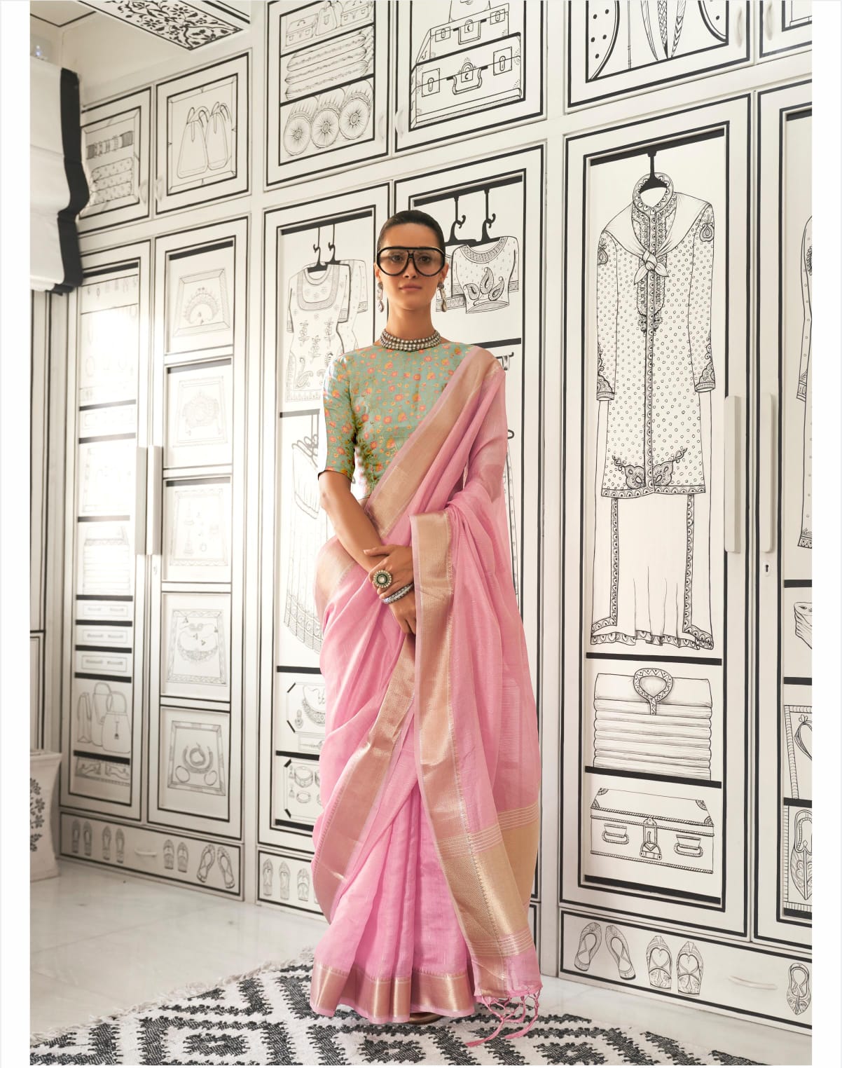 Woman in a pink zari work saree standing indoors against a backdrop of black-and-white fashion illustrations on the walls