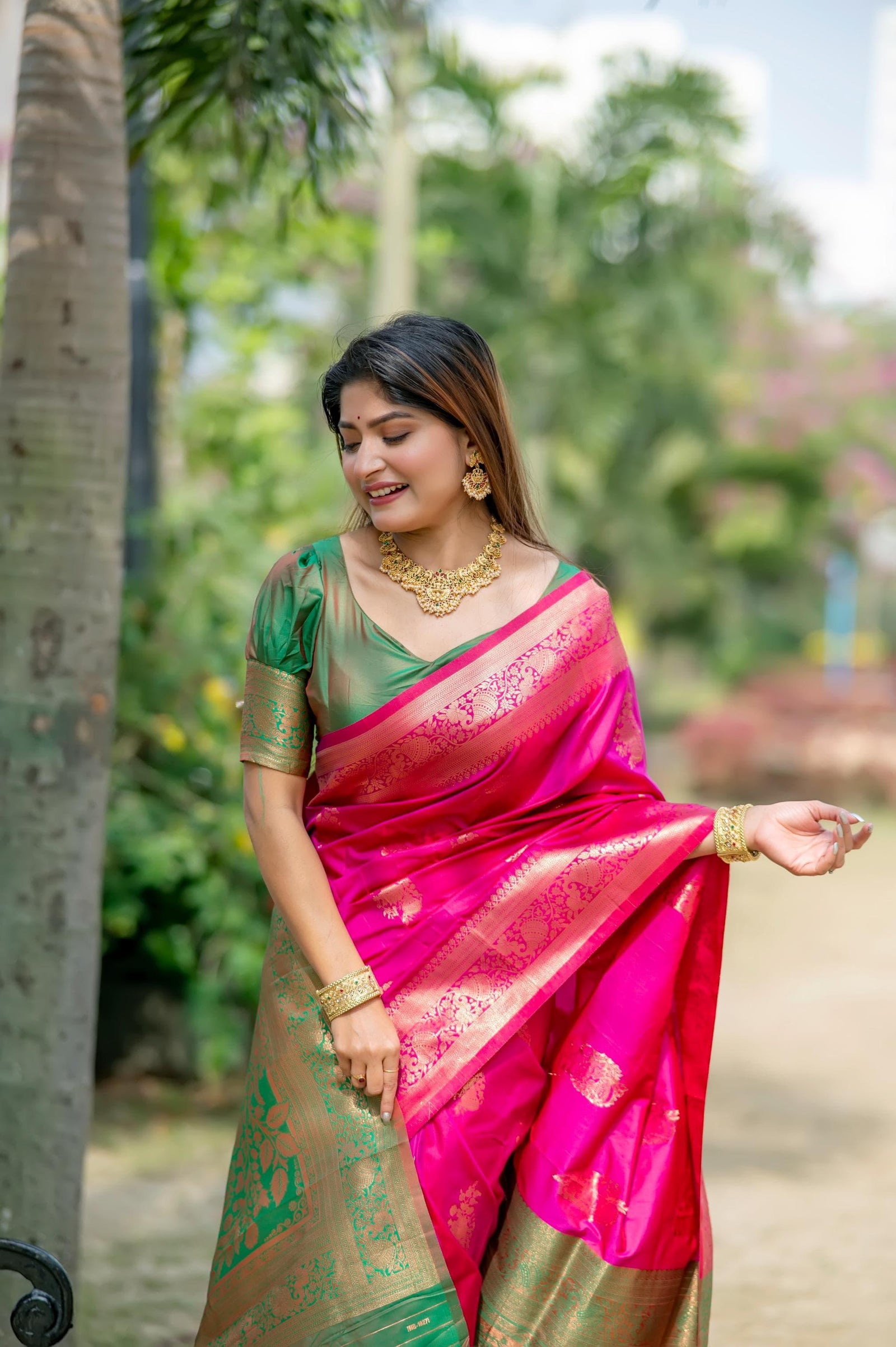 Woman dressed in a pink zari work saree with gold jewelry smiling and posing outdoors in a natural garden setting