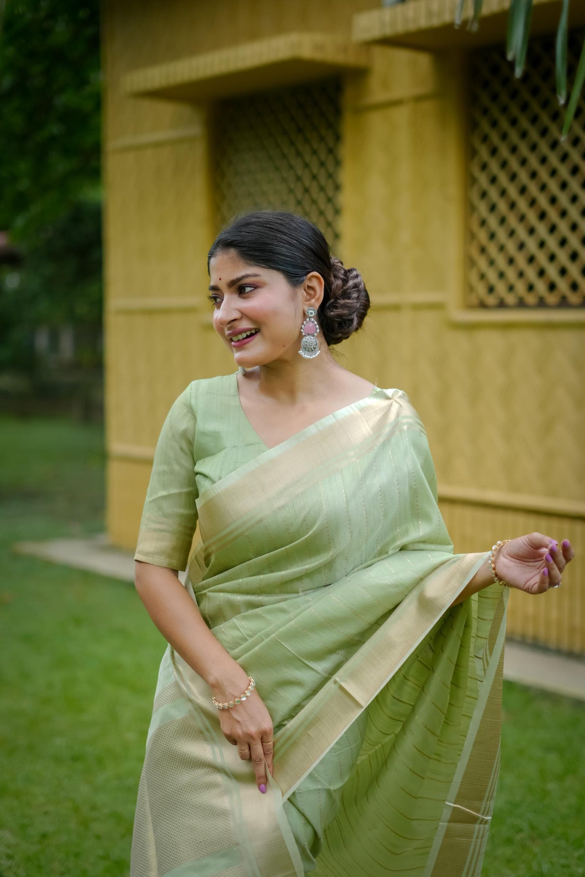 Woman in pista-colored zari work saree standing near a yellow wall with lattice windows and green lawn