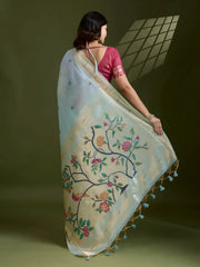 Woman wearing a powder blue silk saree with an off-white floral embroidered pallu and gold zari border, paired with a contrasting magenta blouse and traditional jewelry, elegantly posing in a studio with olive green walls and patterned window light.