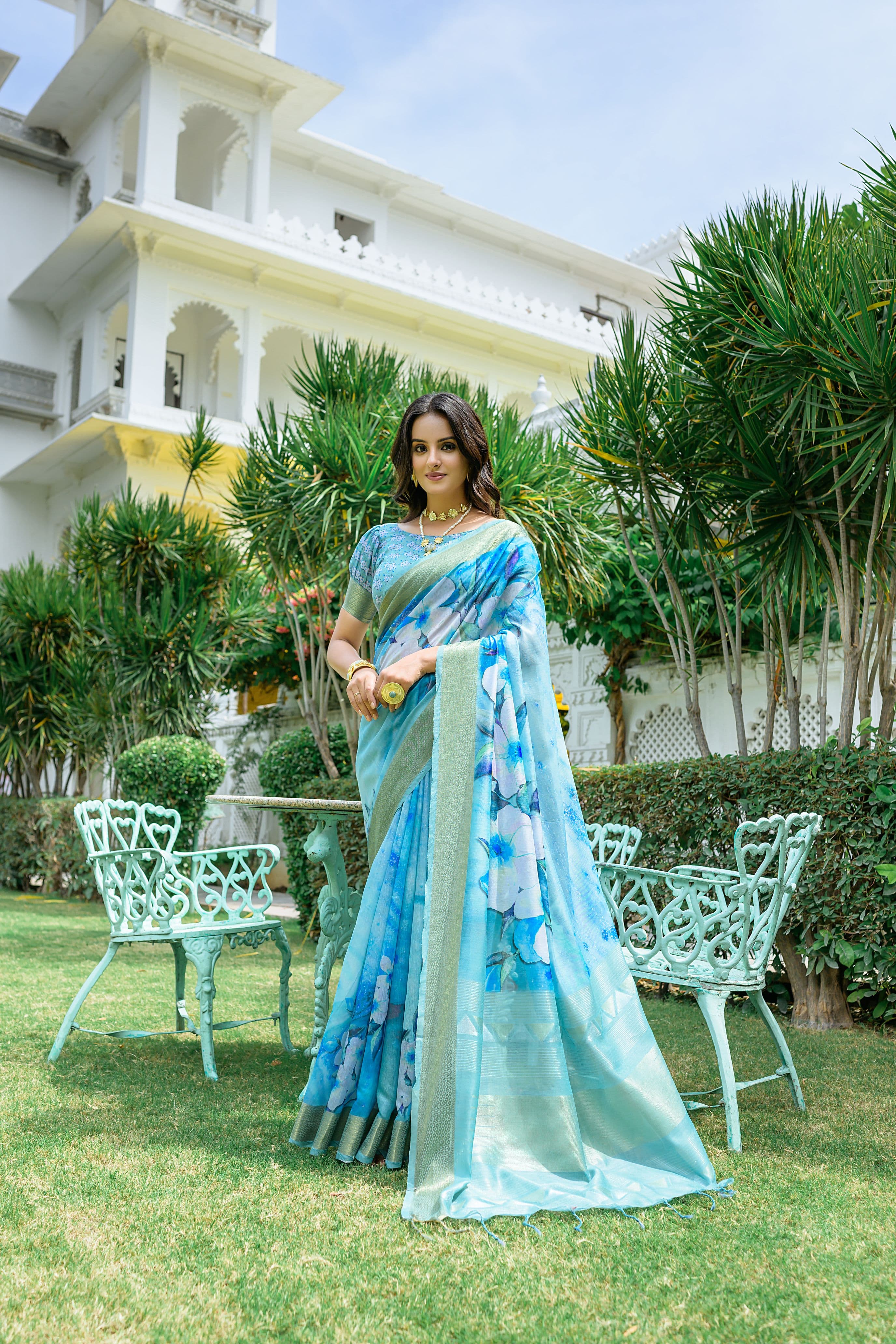Woman wearing powder blue zari work saree with floral prints and golden border in a royal garden setting