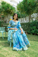 Woman wearing powder blue zari work saree with floral prints and golden border in a royal garden setting