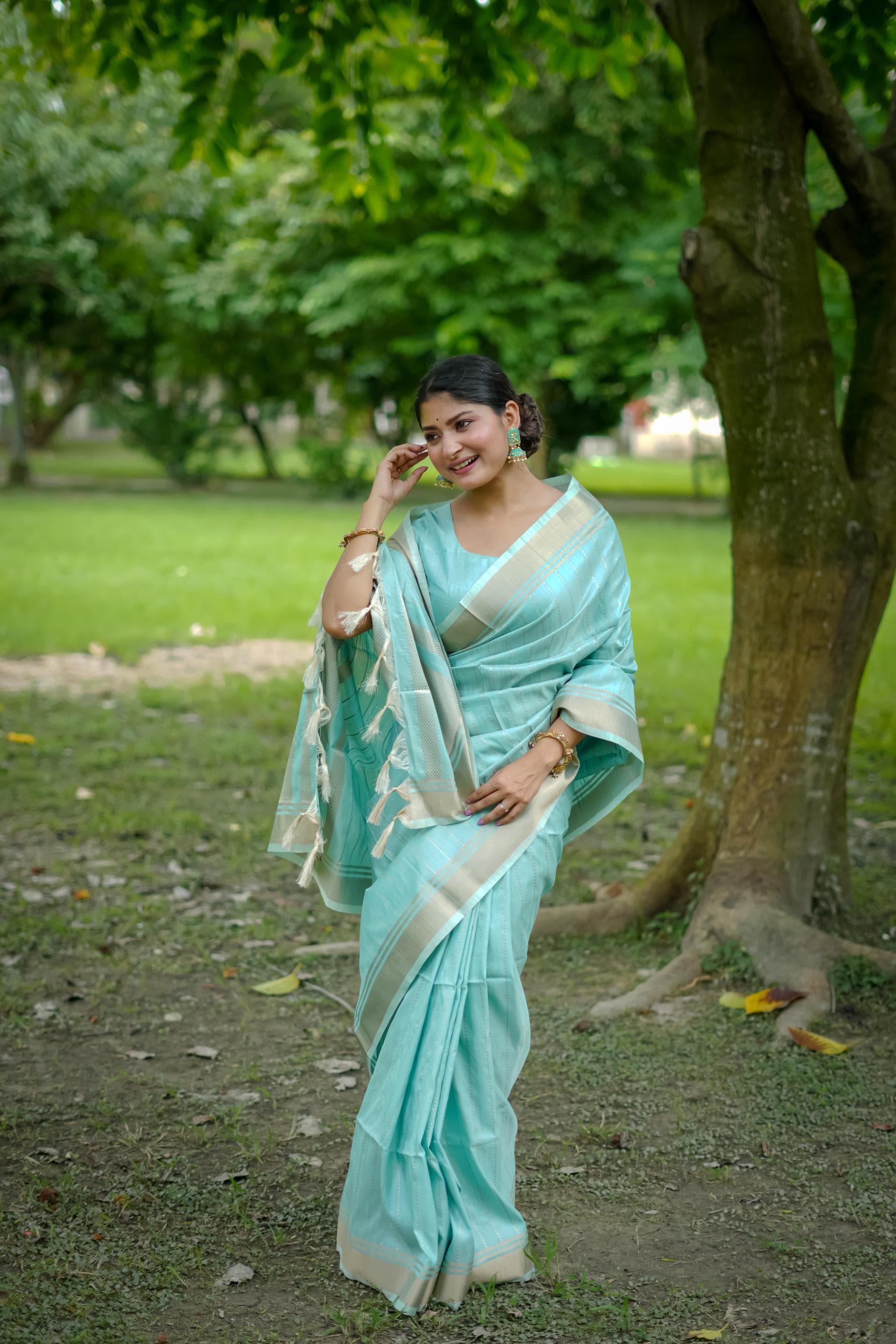 Woman in powder blue zari work saree standing in a lush green park near a tree