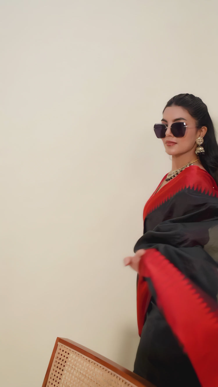 A woman models a striking Black Raw Silk Saree featuring a wide, vibrant deep red border with traditional temple weaving. The saree is cinched with a belt. She wears a red blouse, a metallic choker necklace, and large sunglasses, posing indoors.