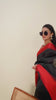 A woman models a striking Black Raw Silk Saree featuring a wide, vibrant deep red border with traditional temple weaving. The saree is cinched with a belt. She wears a red blouse, a metallic choker necklace, and large sunglasses, posing indoors.