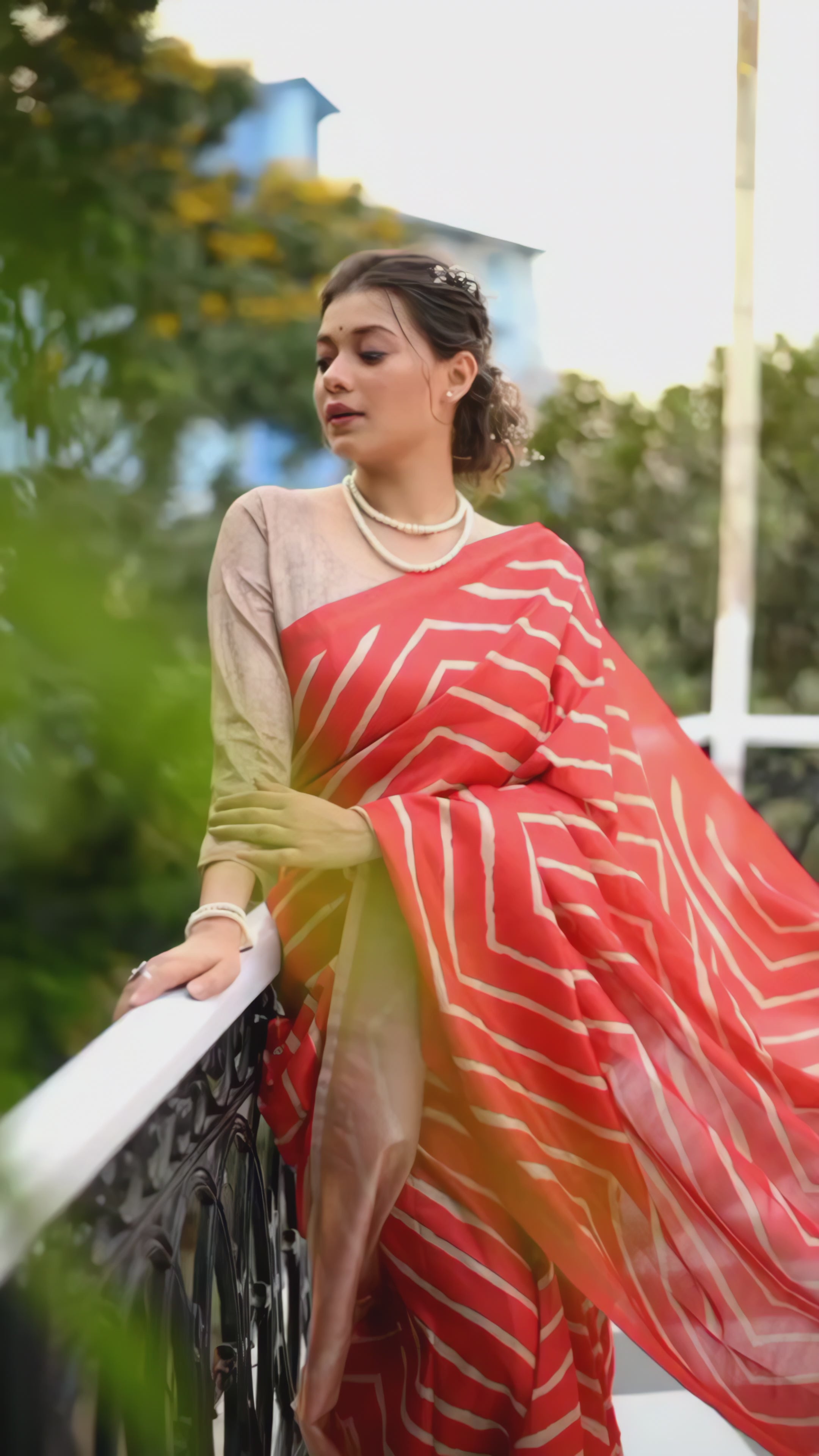 A woman models a vibrant Hot Pink Saree with a bold off-white chevron/striped pattern. The pallu is light beige with a dark border, featuring an elaborate printed motif of colorful flowers. She wears a pearl necklace and a large yellow flower hair accessory, posing indoors.