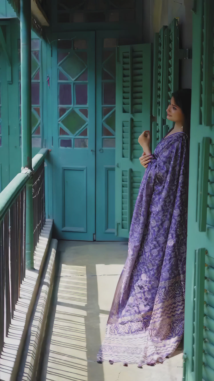A woman models an elegant Deep Purple Tussar Silk Saree. The saree features dense all-over floral embroidery/weaving in a light metallic thread, framed by a contrasting metallic border. She wears a matching purple blouse and accessorizes with heavy oxidized silver jhumka earrings and bangles, posing indoors next to a teal door.