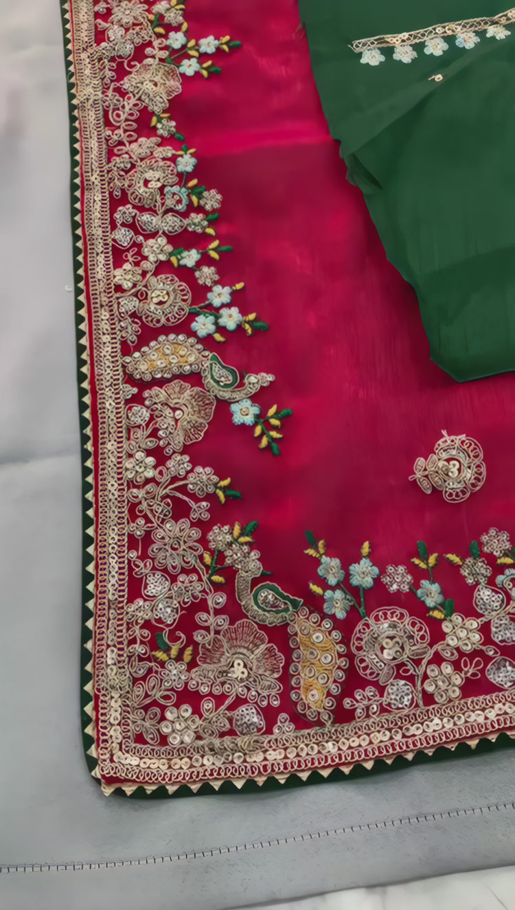 A woman models a vibrant Dark Pink Chiffon Silk Saree with scattered embellishments. The saree has a wide, intricate silver embroidery border. She wears a heavily embroidered dark green blouse and accessorizes with silver jewelry, posing indoors.