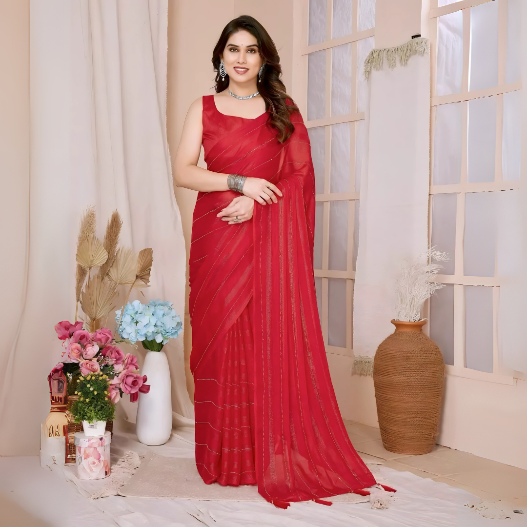 A woman models a vibrant Crimson Red Rimzim Silk Saree. The sheer fabric features prominent vertical stripes and scattered embellishments of fine metallic (silver) threadwork. She wears a matching sleeveless red blouse and accessorizes with chunky oxidized silver jewelry, including a layered necklace and a wide cuff bangle, posing indoors.