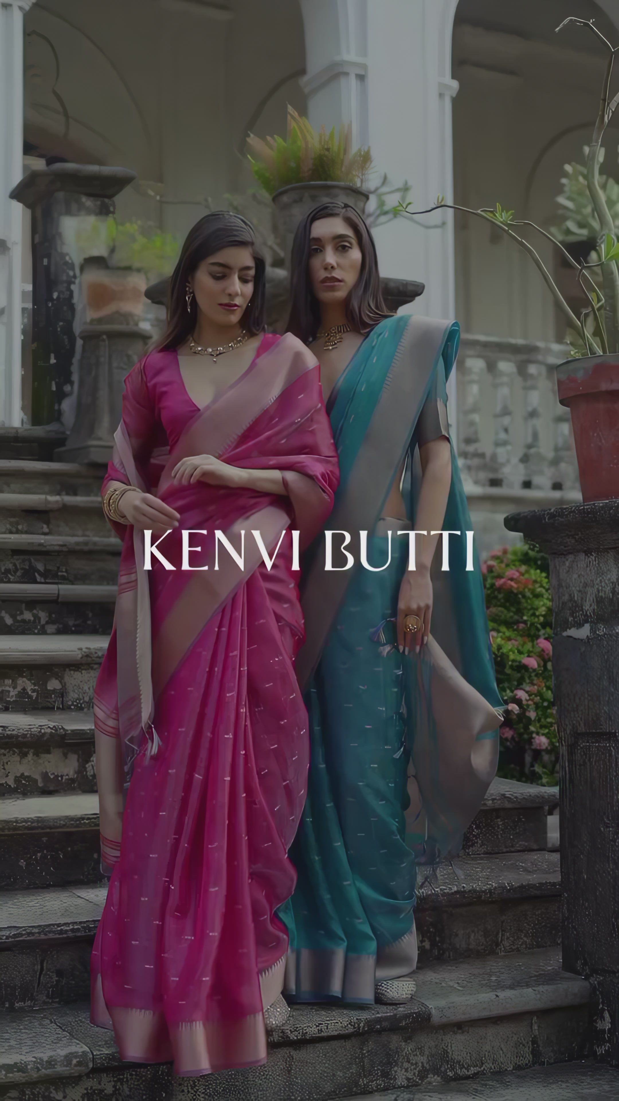 A woman models a stunning Coral Red Soft Silk Saree. The sheer saree features subtle metallic motifs and a wide, contrasting metallic gold/bronze woven border and pallu. She wears a matching red short-sleeve blouse and delicate gold jewelry, posing gracefully on an ornate stone staircase.