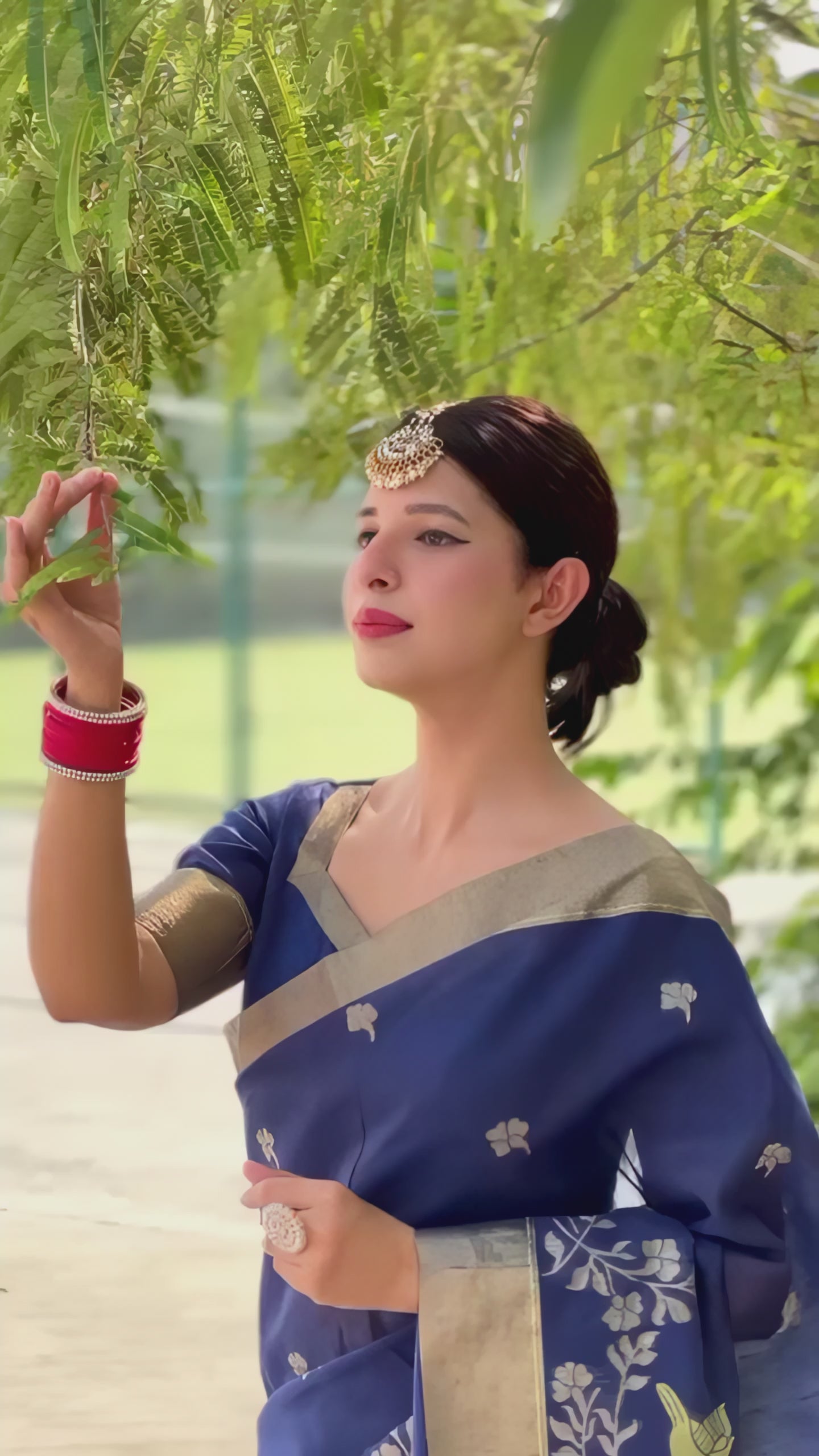 A woman models a vibrant Navy Blue Linen Silk Saree with delicate gold/silver Zari woven motifs and a wide metallic silver border. She wears a beige blouse, red and white bangles, and a traditional headpiece, posing outdoors under a tree.