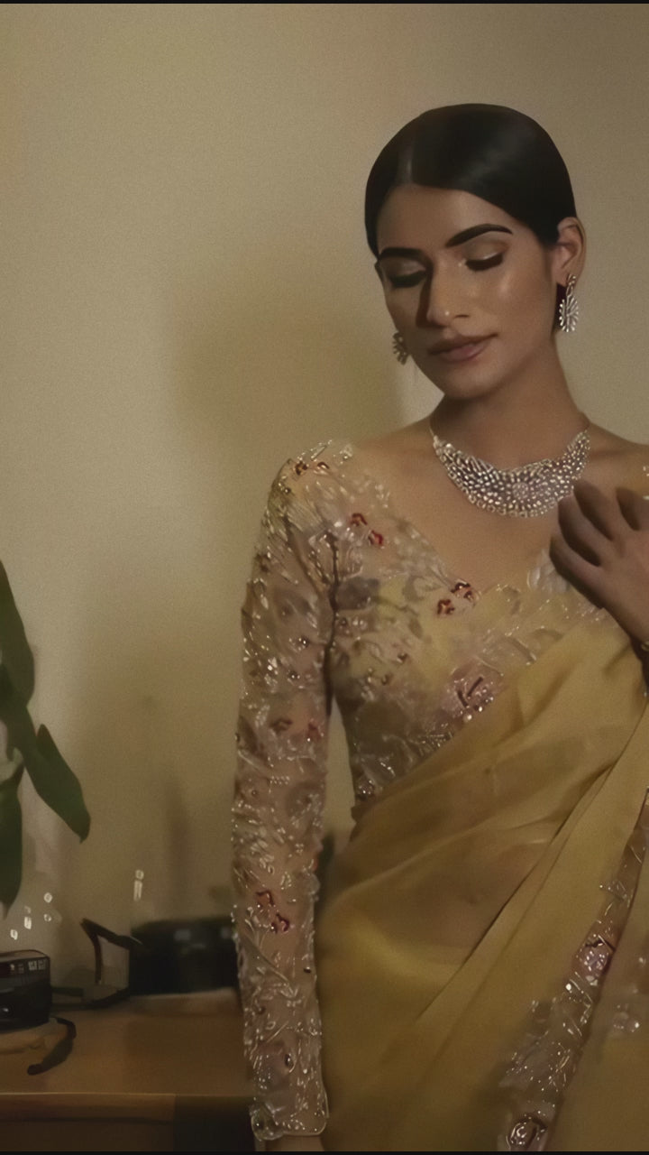 A woman models a striking Golden Yellow Soft Organza Silk Saree. The saree features delicate thread and sequin floral embroidery along the border. She wears a matching long-sleeve, heavily embroidered blouse and accessorizes with silver/diamond jewelry.