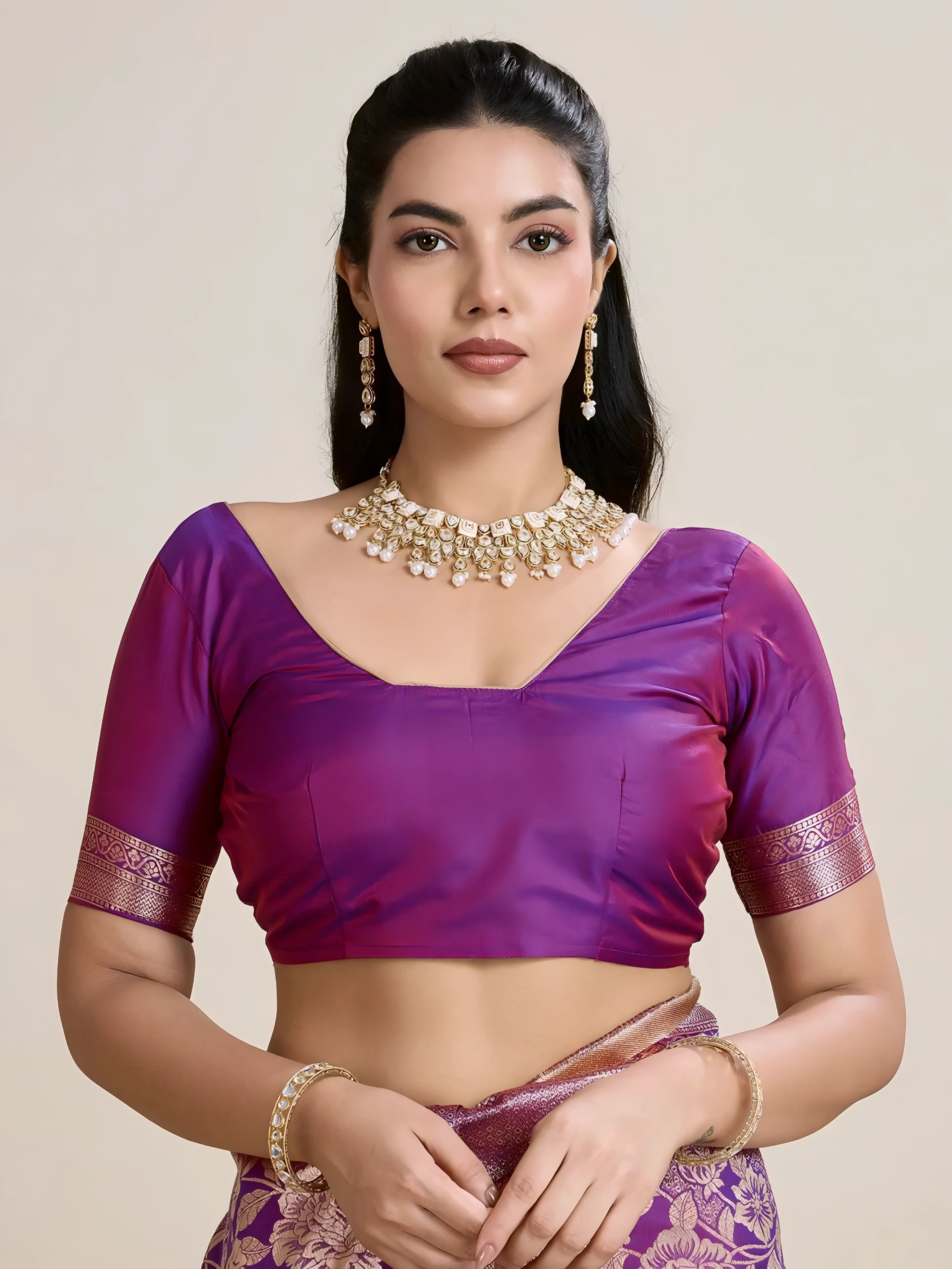 A woman models a luxurious Deep Violet Soft Kanjivaram Silk Saree. The saree features striking large floral and scattered motifs woven in metallic rose gold Zari on the body and wide pallu. She wears a matching short-sleeve violet blouse and accessorizes with a heavy gold Kundan choker necklace and earrings.