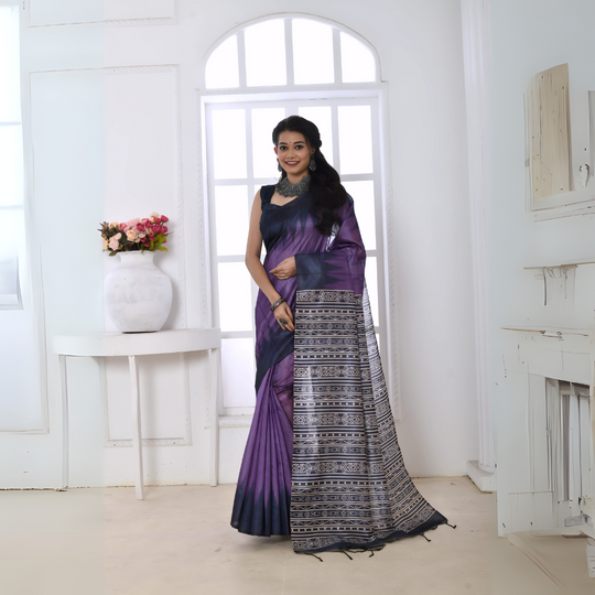 A woman models a striking Purple Soft Tussar Silk Saree. The saree features a color-block transition between purple and deep navy/black. The pallu has a wide panel of dense, grey and black geometric Ikat-style print. She wears a dark sleeveless blouse and chunky oxidized silver jewelry.