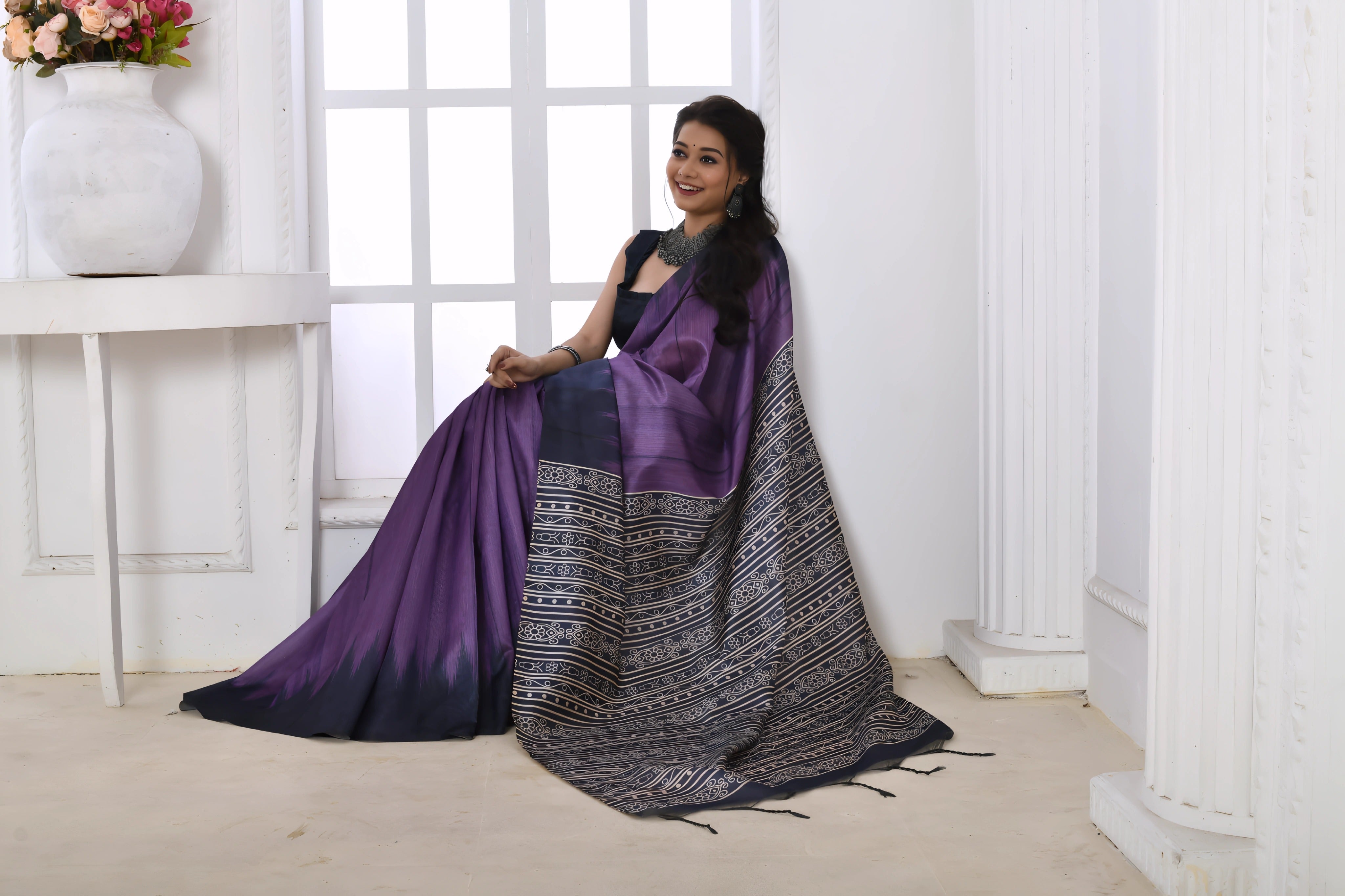 A woman models a striking Purple Soft Tussar Silk Saree. The saree features a color-block transition between purple and deep navy/black. The pallu has a wide panel of dense, grey and black geometric Ikat-style print. She wears a dark sleeveless blouse and chunky oxidized silver jewelry.