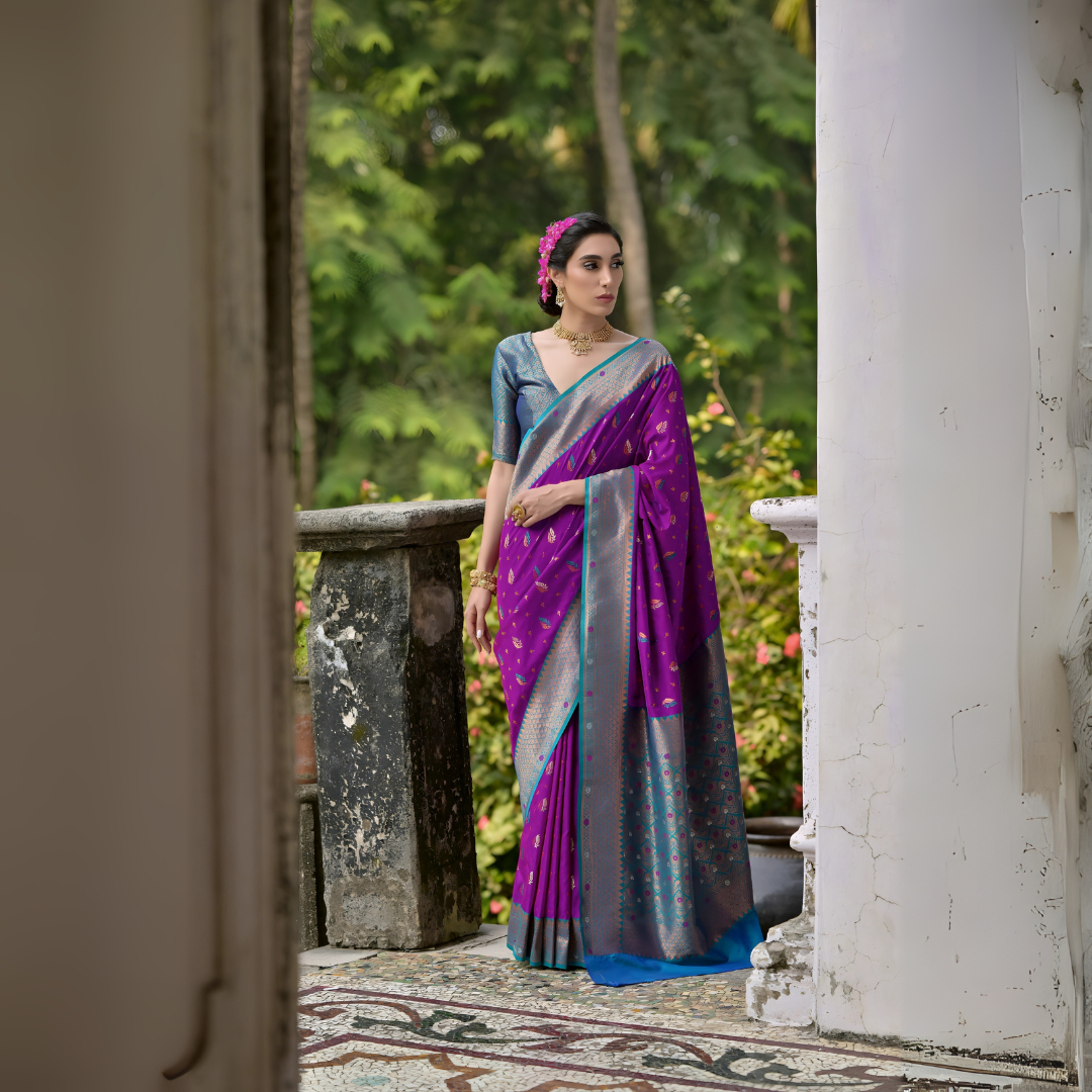 A woman models a vibrant Royal Purple Banarasi Silk Saree with scattered gold and multi-colored Zari motifs. The wide border and pallu contrast dramatically in Rama Blue/Teal with dense traditional gold weaving. She wears a grey-blue blouse and gold Kundan jewelry, posing on a tiled outdoor veranda.