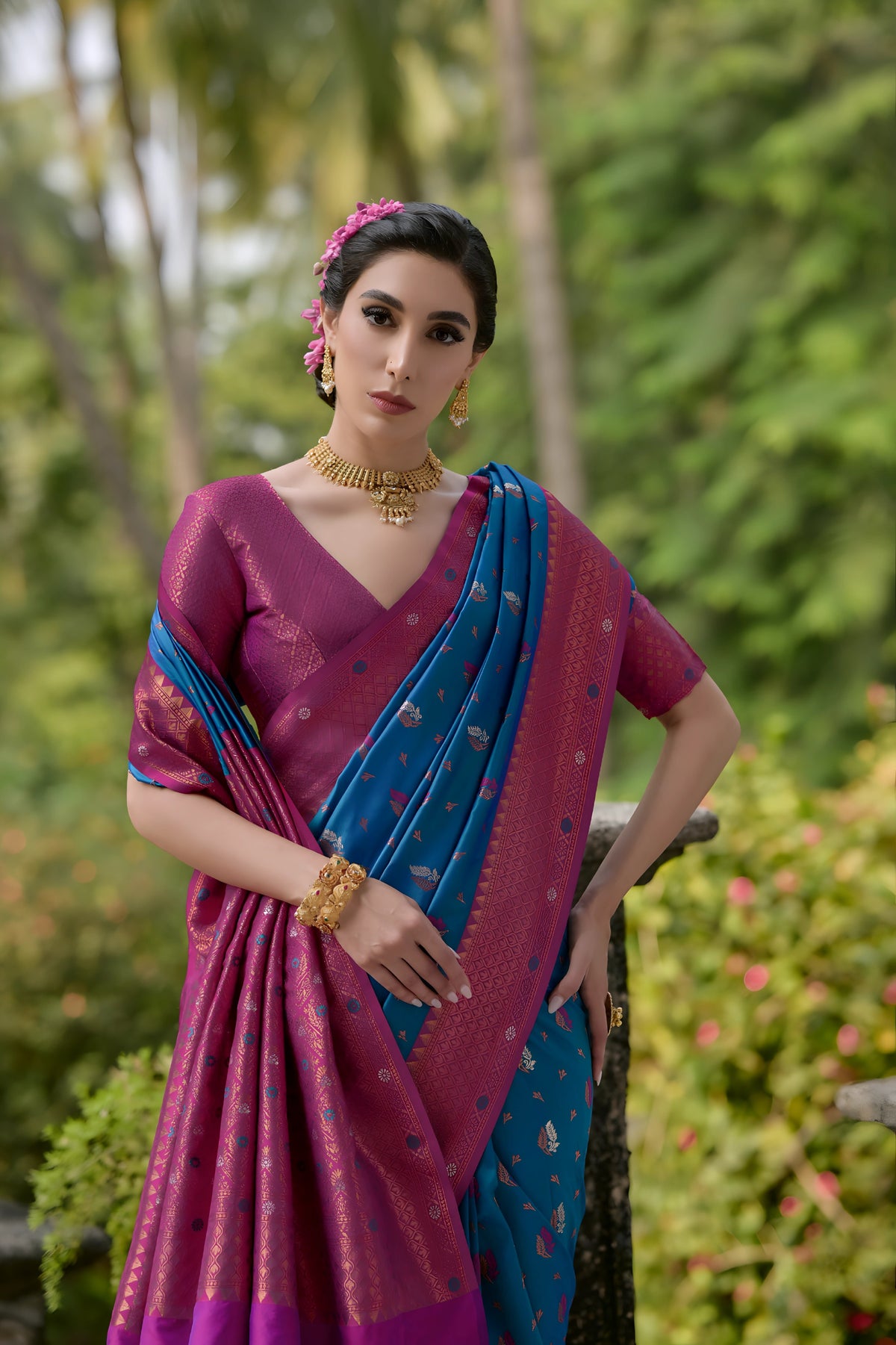 A woman models a vibrant Royal Purple Banarasi Silk Saree with scattered gold and multi-colored Zari motifs. The wide border and pallu contrast dramatically in Rama Blue/Teal with dense traditional gold weaving. She wears a grey-blue blouse and gold Kundan jewelry, posing on a tiled outdoor veranda.
