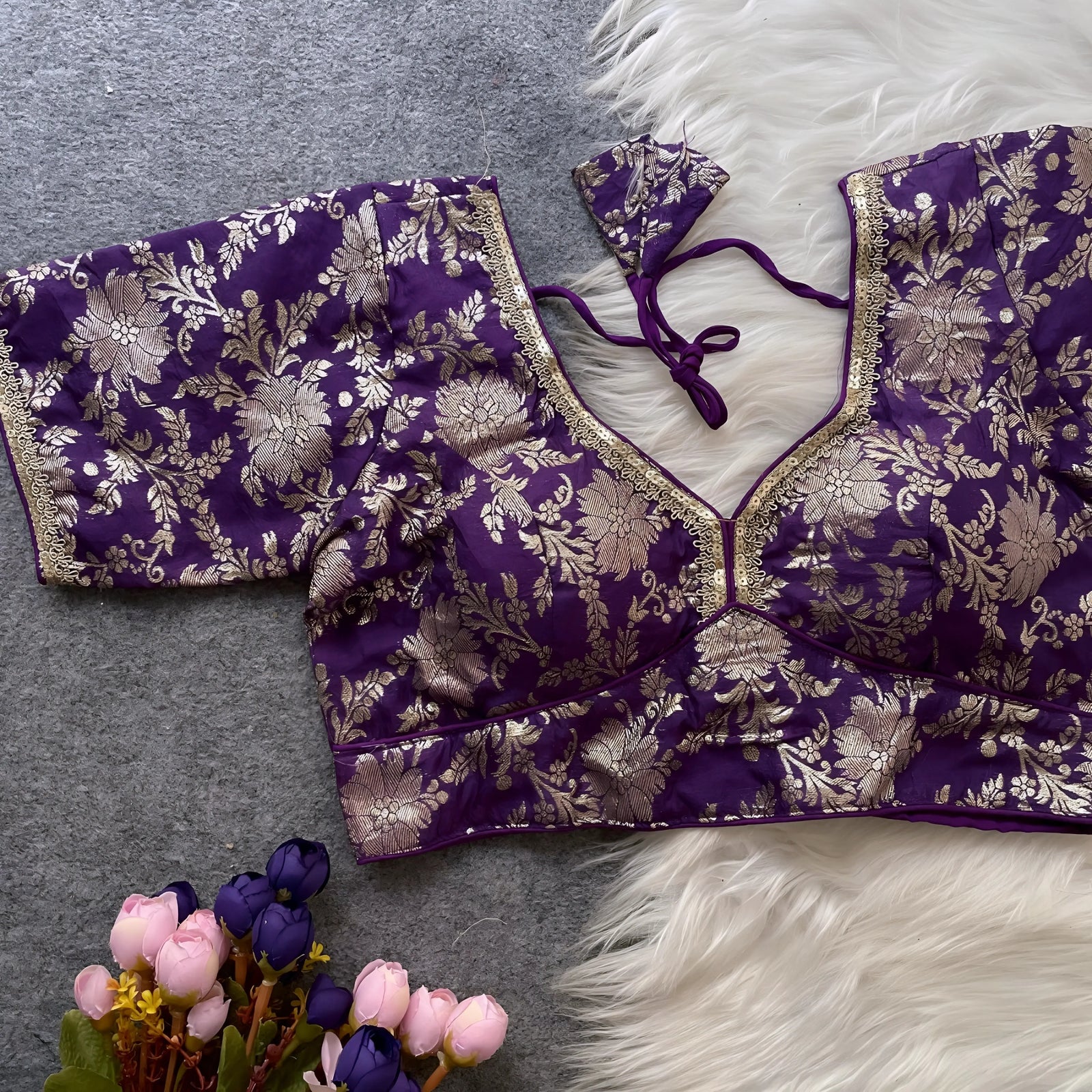 Purple brocade blouse featuring intricate gold zari floral patterns and decorative trim on the neckline and sleeves, displayed flat on a grey surface with a white faux fur accent and a bunch of artificial purple and pink flowers for an elegant, festive look.