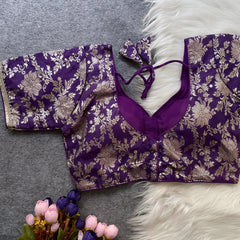 Purple brocade blouse featuring intricate gold zari floral patterns and decorative trim on the neckline and sleeves, displayed flat on a grey surface with a white faux fur accent and a bunch of artificial purple and pink flowers for an elegant, festive look.
